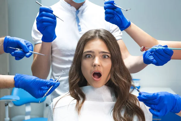 Joven morena sorprendida paciente sentada en silla de dentista rodeada de manos en guantes azules con instrumentos médicos — Foto de Stock