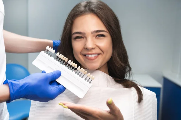 Uśmiechnięta brunetka kobieta pacjenta o appiontment w klinice dentystycznej podnoszenia cieniu za pomocą szkliwa zębów Skala posiadanych przez dentystę w niebieskie rękawice — Zdjęcie stockowe