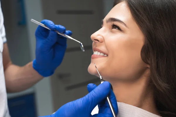 Sonriente morena feliz paciente examinada por dentista en guantes azules usando espejo dental y escalador sentado medio turno en silla dental — Foto de Stock