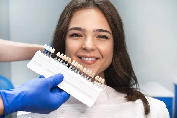 Leende lycklig brunett kvinna patient med appiontelse i Dental Clinic plocka upp skugga med hjälp av tandemaljen skala innehas av tandläkare i blå handskar — Stockfoto