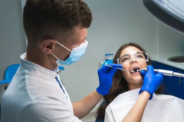 Dentysta w maski medycznej napełniania pacjentów kanał korzeniowy, podczas gdy ona jest w fotelu stomatologicznym noszenie okulary pod lampą medyczną w klinice — Zdjęcie stockowe