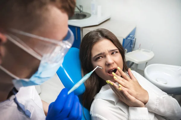 Belle jeune femme patiente brune effrayée ayant un examen au cabinet dentaire par un beau dentiste en masque et gants bleus — Photo