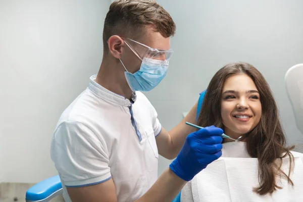 Jeune jolie patiente brune examinée par un dentiste en gants bleus masque médical et lunettes de sécurité à l'aide d'un miroir dentaire dans une clinique dentaire — Photo