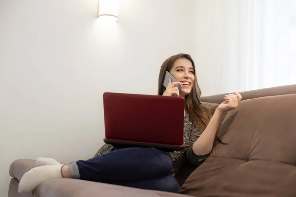 Young beautiful smiling brunette girl sitting on the sofa talking on the phone and watching movie on laptop — Stock Photo, Image