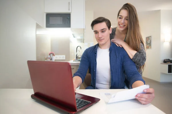 Jonge knappe man werken in zijn laptop van huis studeren documenten zijn mooie vrouw glimlachend staande achter hem — Stockfoto