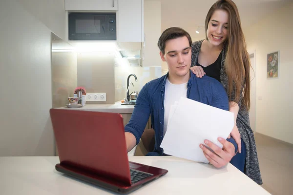 Feliz pareja - joven hombre guapo trabajando en su computadora portátil desde casa estudiando documentos su hermosa esposa sonriendo de pie detrás de él — Foto de Stock