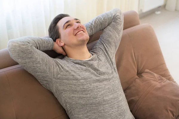 Молодой красивый улыбающийся мужчина, сидящий на диване с руками за головой, чувствуя себя расслабленным и спокойным — стоковое фото