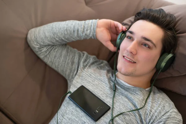 Joven guapo relajado hombre acostado en el sofá escuchando música en los auriculares desde el teléfono inteligente sensación de paz —  Fotos de Stock