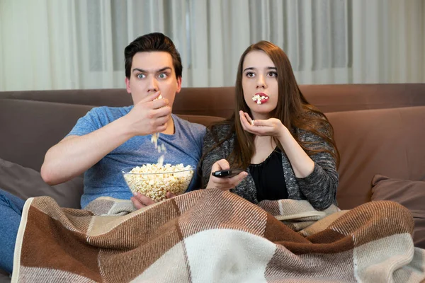 young couple watching movie on the sofa handsome man holding plate with popcorn attractive woman holding remote control both looking scared