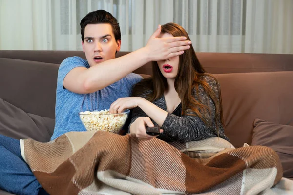 Joven pareja viendo película en el sofá comiendo palomitas guapo hombre cubriendo sus esposas ojos con su mano ambos buscando asustado —  Fotos de Stock