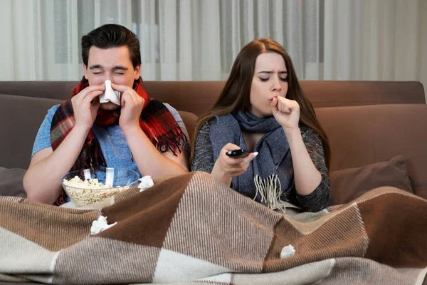 Jovem casal assistindo filme no sofá bonito homem soprando o nariz e mulher atraente tossir ambos vestindo cachecóis olhando doente — Fotografia de Stock