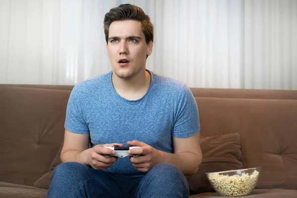 Joven guapo hombre jugando video juego sentado en cómodo sofá buscando fascinado —  Fotos de Stock