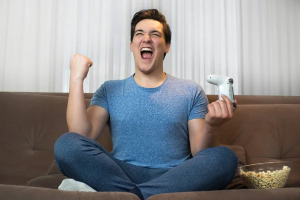 Joven guapo hombre jugando videojuego sentado en cómodo sofá sosteniendo joystick buscando feliz misión completada con éxito —  Fotos de Stock