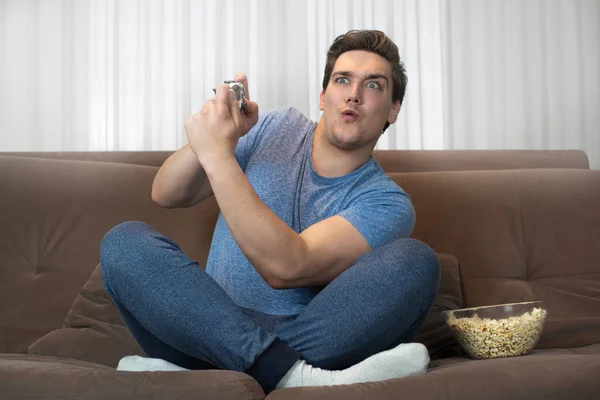 Joven guapo hombre jugando videojuego sentado en cómodo sofá sosteniendo joystick con ambas manos tener momentos tensos en completar la misión —  Fotos de Stock