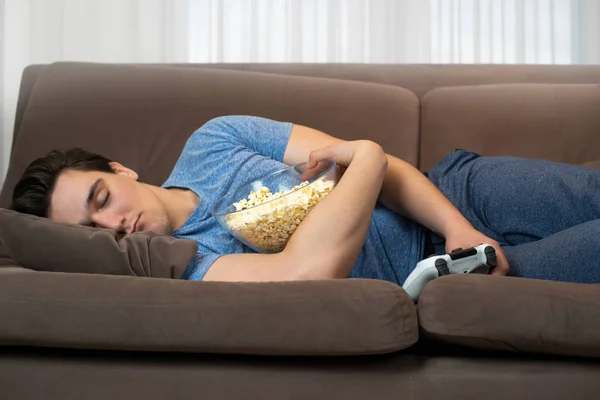 Молодой красивый мужчина засыпает после игры в увлекательную видеоигру, держа в одной руке джойстик, а в другой попкорн. — стоковое фото