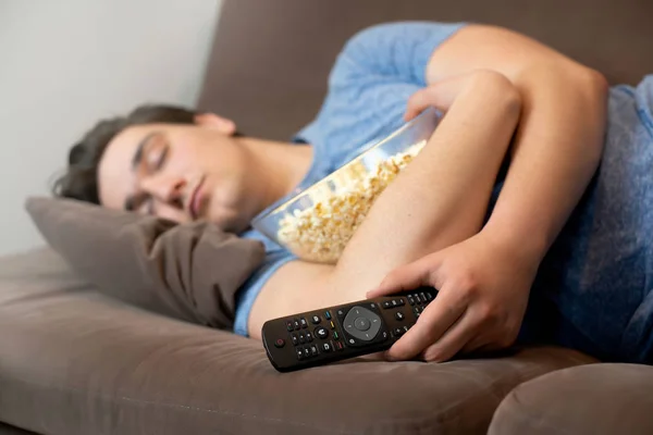 Молодой красивый мужчина засыпает после просмотра захватывающего фильма с пультом дистанционного управления в одной руке и попкорном в другой — стоковое фото