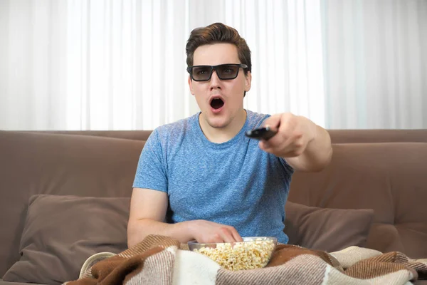 Joven sorprendido hombre usando gafas de sol viendo película comer palomitas de maíz clic control remoto —  Fotos de Stock