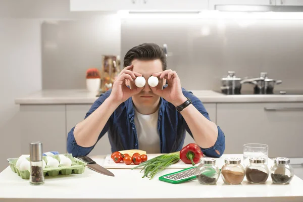 Joven hombre guapo sentado en la cocina preparando el desayuno cubre sus ojos con huevos frescos que se divierten mucho — Foto de Stock