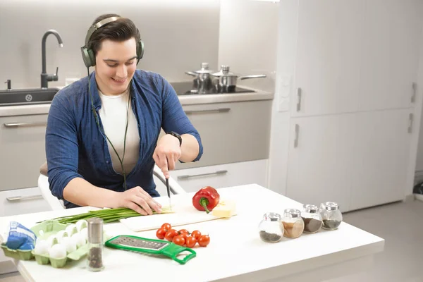 Ung stilig man bär hörlurar håller kniv i ena handen med mycket roligt skära purjolök förbereder frukost i ljust kök — Stockfoto