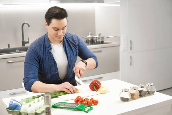 Ung stilig man hållai kniv i ena handen med mycket roligt skära färska purjolök förbereder frukost i ljust kök — Stockfoto