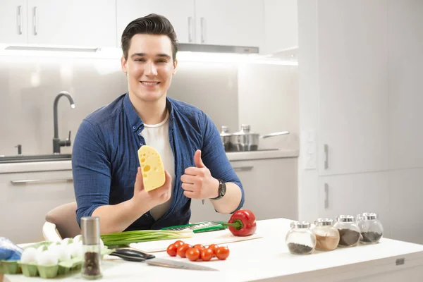 Ung stilig leende man sitter i ljust kök förbereder frukost som håller färsk ost i ena handen och visar OK skylt — Stockfoto