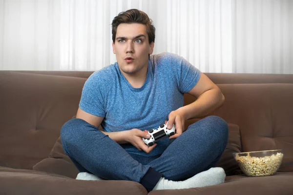 Joven guapo hombre jugando videojuego sentado en cómodo sofá sosteniendo joystick se ve muy concentrado en la misión —  Fotos de Stock