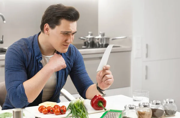 Ung stilig man sitter i ljust kök förbereder frukost ser förbryllad med recepie i handen — Stockfoto