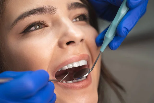 Sonriente hermosa mujer morena feliz paciente examinado por el dentista en guantes azules usando espejo dental y escalador sentado en la clínica dental de cerca — Foto de Stock