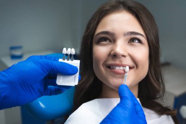 dentist in blue medical gloves applying sample from tooth enamel scale to happy woman patient teeth to pick up right shade for teeth bleaching procedure clipart