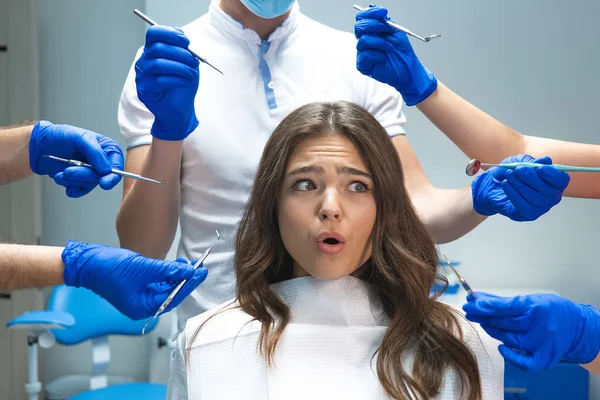 Scared young brunette woman patient sitting in dentist chair surrounded by hands in blue gloves with medical instruments — Stock Photo, Image