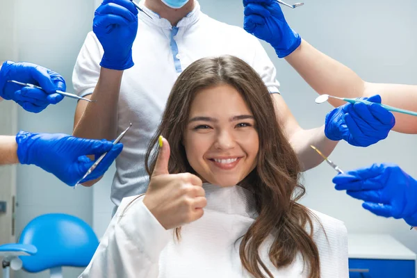 Heureuse jeune femme brune patiente assise sur une chaise de dentiste entourée de mains en gants bleus avec instruments médicaux et dentiste masqué debout derrière — Photo