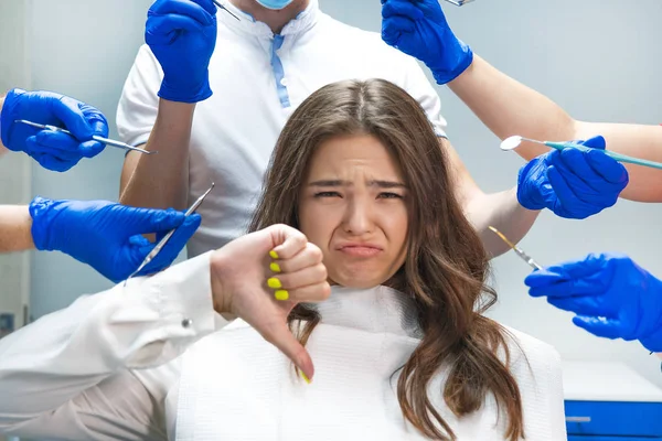 Rozrušená mladá brunetka žena, která sedí v zubařské židli obklopená rukama v modrých rukavicích s lékařskými nástroji a zubařem v masce, stojící za — Stock fotografie