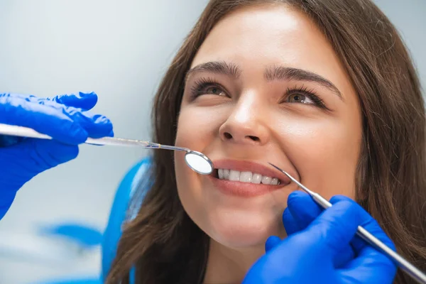 Uśmiechnięty szczęśliwy brunetka kobieta pacjent badane przez dentystę w niebieskie rękawiczki za pomocą lustro dentystyczne i skaler siedzi w klinice dentystycznej — Zdjęcie stockowe