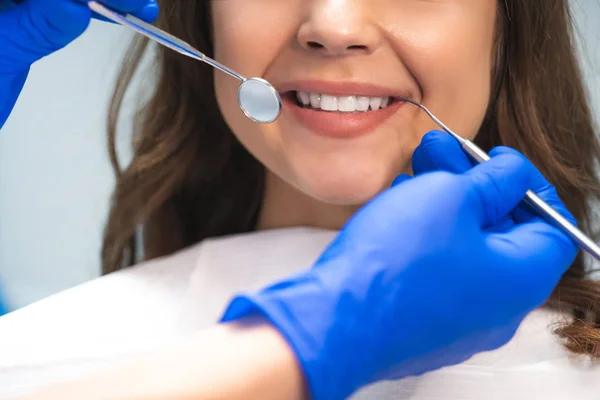 Uśmiechnięta brunetka kobieta pacjenta badane przez dentystę w niebieskie rękawiczki za pomocą lustro dentystyczne i skaler siedzi w klinice dentystycznej — Zdjęcie stockowe
