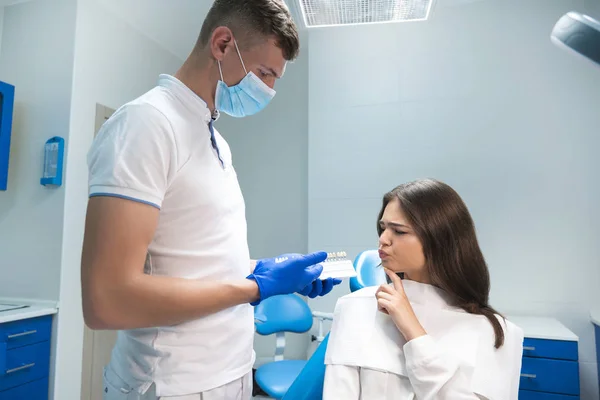 Zubař na lékařské masce a s modrými rukavicemi ukazující na zubovou stupnici k usměvavé ženě pacient na bělení zubů na stomatologické klinice — Stock fotografie