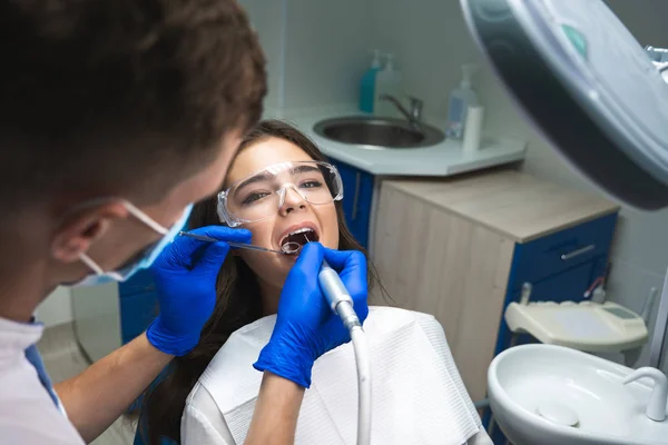 Dentysta w masce napełniania kanału korzeniowego pacjentów, podczas gdy ona leży na fotelu dentystycznym noszenia okularów pod lampą medyczną w klinice — Zdjęcie stockowe