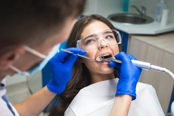 Dentysta w masce napełniania kanału korzeniowego pacjentów, podczas gdy ona leży na krześle dentystycznym w okularach ochronnych pod lampą medyczną w klinice — Zdjęcie stockowe