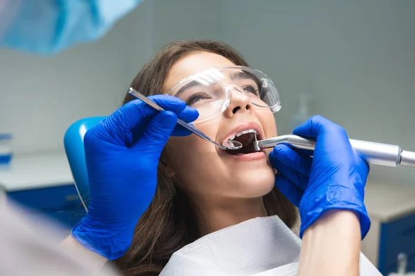 Dentysta w masce napełniania kanału korzeniowego pacjentów, podczas gdy ona leży na fotelu dentystycznym noszenie okularów pod lampą medyczną w gabinecie stomatologicznym — Zdjęcie stockowe