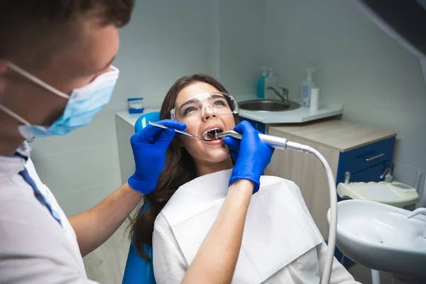 Dentysta w maski medycznej napełniania pacjentów kanał korzeniowy, podczas gdy ona leży na fotelu dentystycznym noszenie okulary pod lampą medyczną w gabinecie stomatologicznym — Zdjęcie stockowe