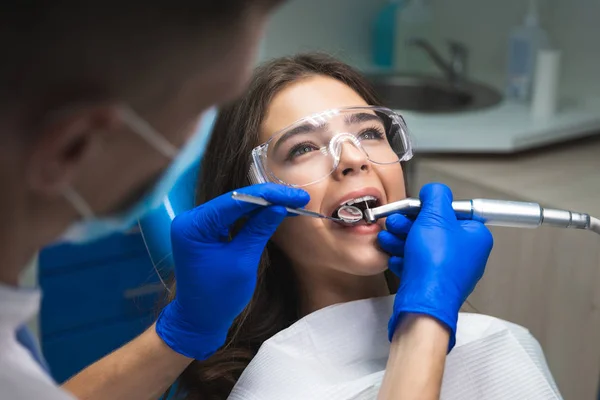 Młoda brunetka kobieta pacjenta badane przez dentystę w niebieskie rękawiczki i maski medycznej za pomocą lustro dentystyczne i skaler siedzi w klinice dentystycznej — Zdjęcie stockowe