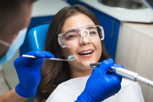 Sonriente morena feliz paciente examinada por el dentista en guantes azules y máscara médica usando espejo dental y escalador sentado en la clínica dental — Foto de Stock