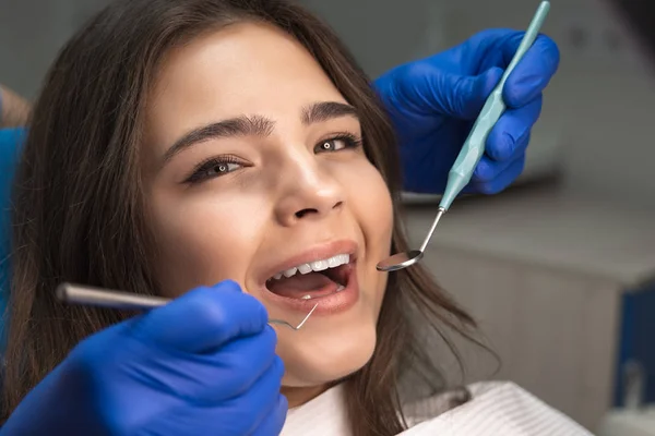 Uśmiechnięty atrakcyjny szczęśliwy kobieta pacjent zbadane przez dentystę w niebieskie rękawiczki za pomocą lustro dentystyczne i skaler siedzi w klinice dentystycznej zbliżenie — Zdjęcie stockowe