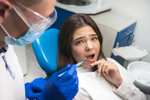 Belle jeune femme patiente effrayée ayant examen au cabinet dentaire par beau dentiste en masque et gants bleus semble effrayé — Photo