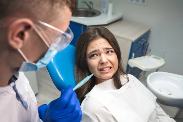 Belle jeune femme brune effrayée patient ayant un examen au cabinet dentaire par un beau dentiste en masque et gants bleus — Photo