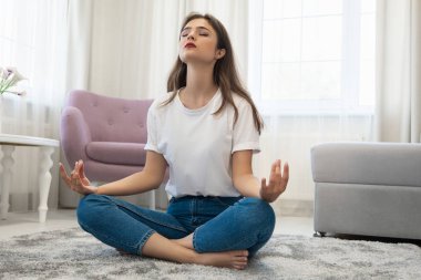 beautiful young brunette woman wearing jeans and white t-shirt sitting on the floor in asana yoga position in bright livingroom looking peaceful clipart