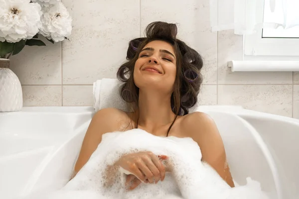 Attrayant jeune femme brune dans les bigoudis de cheveux profiter de son bain avec de la mousse dans la salle de bain lumineuse — Photo
