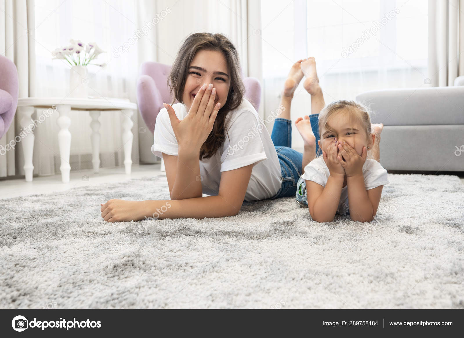Hermosa Joven Morena Mujer Con Lindo Bebe Nina Con Pantalones Vaqueros Y Camisetas Blancas Tumbados En El Suelo En Luminoso Salon Reir Cerrando La Boca Fotografia De Stock C Smiiankaterina Depositphotos