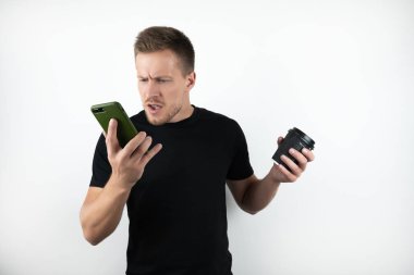 yakışıklı genç sürpriz adam akıllı telefon konuşma siyah t-shirt giyen ve kahve fincan tutan izole beyaz arka plan üzerinde kızgın görünüyor