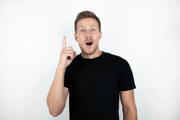 Knappe jonge verrast man dragen zwart t-shirt wijzend met zijn vinger omhoog geïsoleerde witte achtergrond — Stockfoto