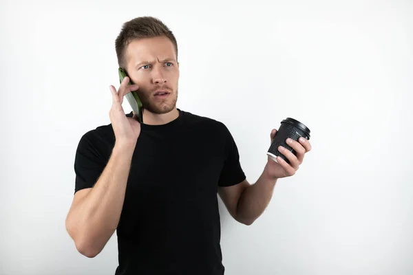 Handsome young surprised man wearing black t-shirt speaking on smartphone and holding cup of coffee on isolated white background — Stock Photo, Image